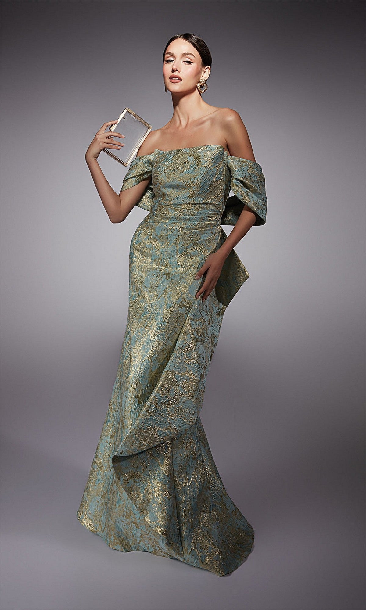 A woman in the Alyce 27750 long formal dress, a metallic jacquard brocade mermaid skirt gown, poses holding a rectangular clutch against a gray studio background.